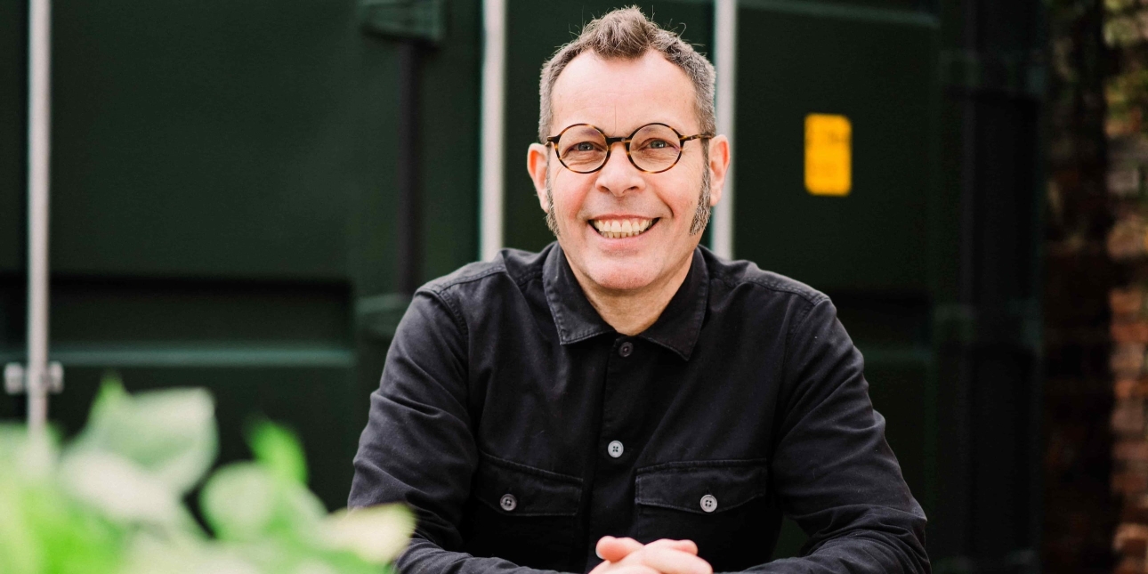 A portrait of a smiling Paul Mackman, a white man with short brown hair and glasses, wearing circular glasses and dark shirt whilst sat at an outdoor table. Behind him is a green shipping metal panel and brickwork